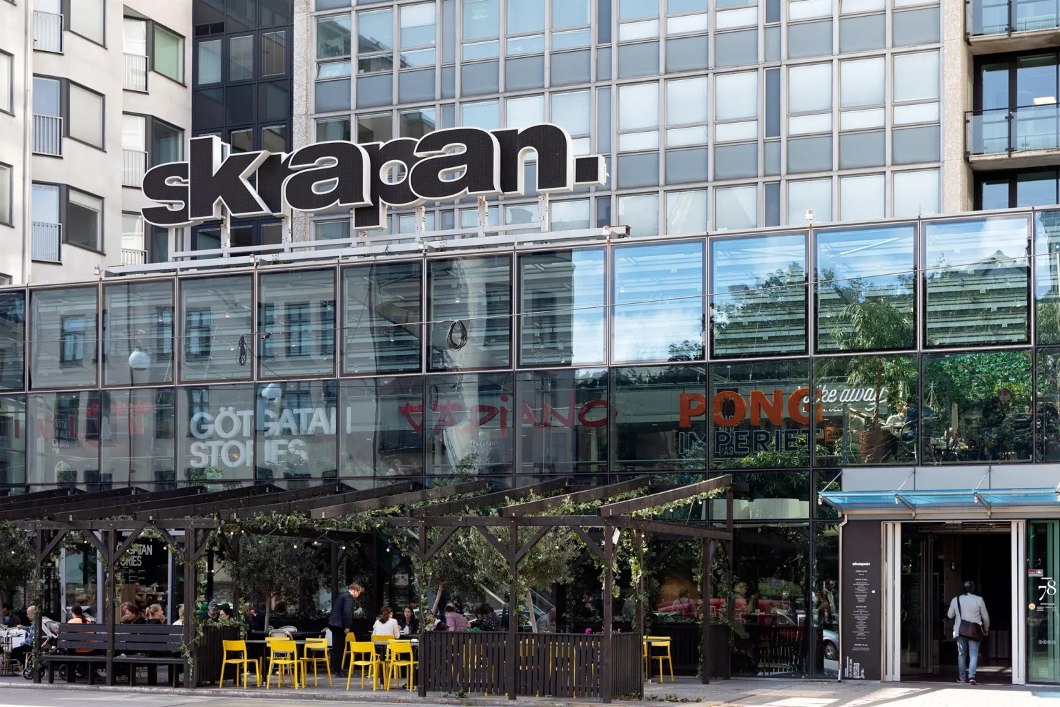 1. En modern byggnad med glasfasad, texten "kapan." syns på fasaden, och en uteservering med gula stolar finns framför.