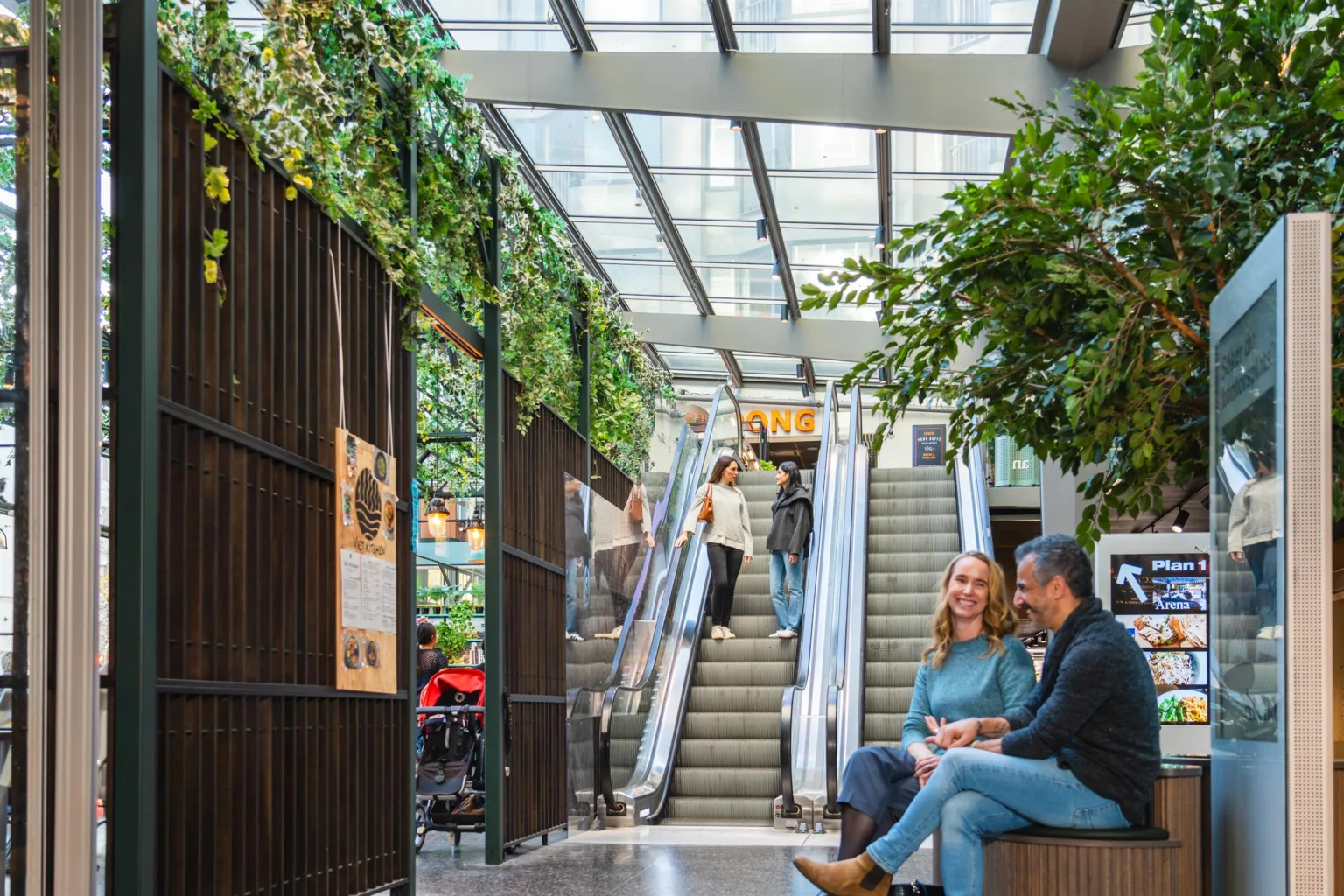 4. Ett inomhuscentrum med gröna växter, rulltrappor och människor som sitter och pratar i en ljus miljö.