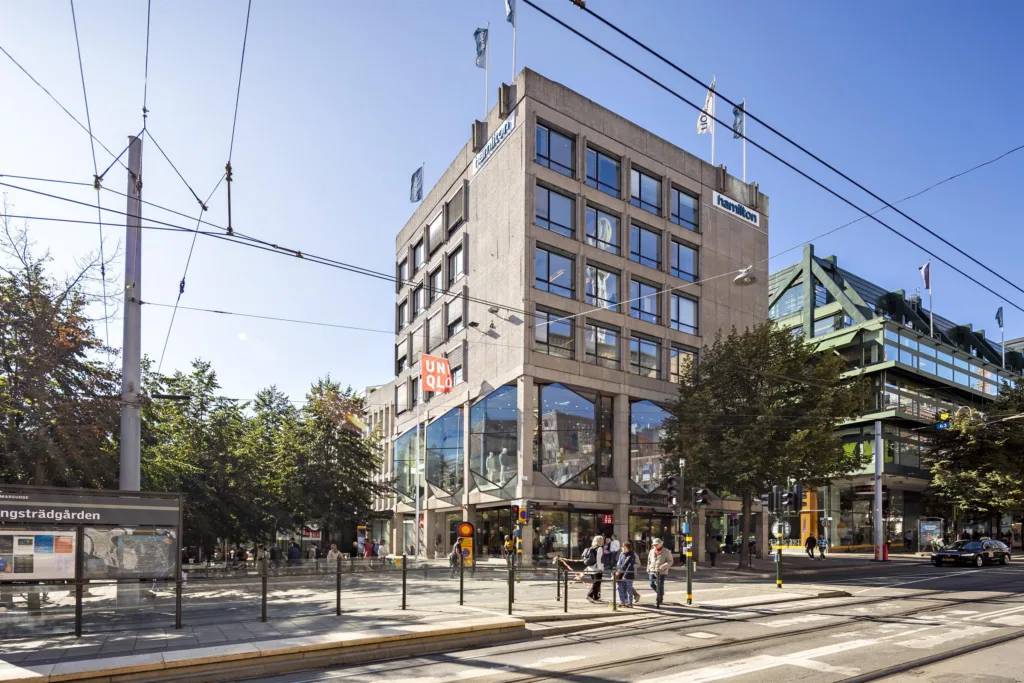 En mindre byggnad med glasfasad och moderna detaljer, omgiven av träd, människor och spårvagnslinjer.
