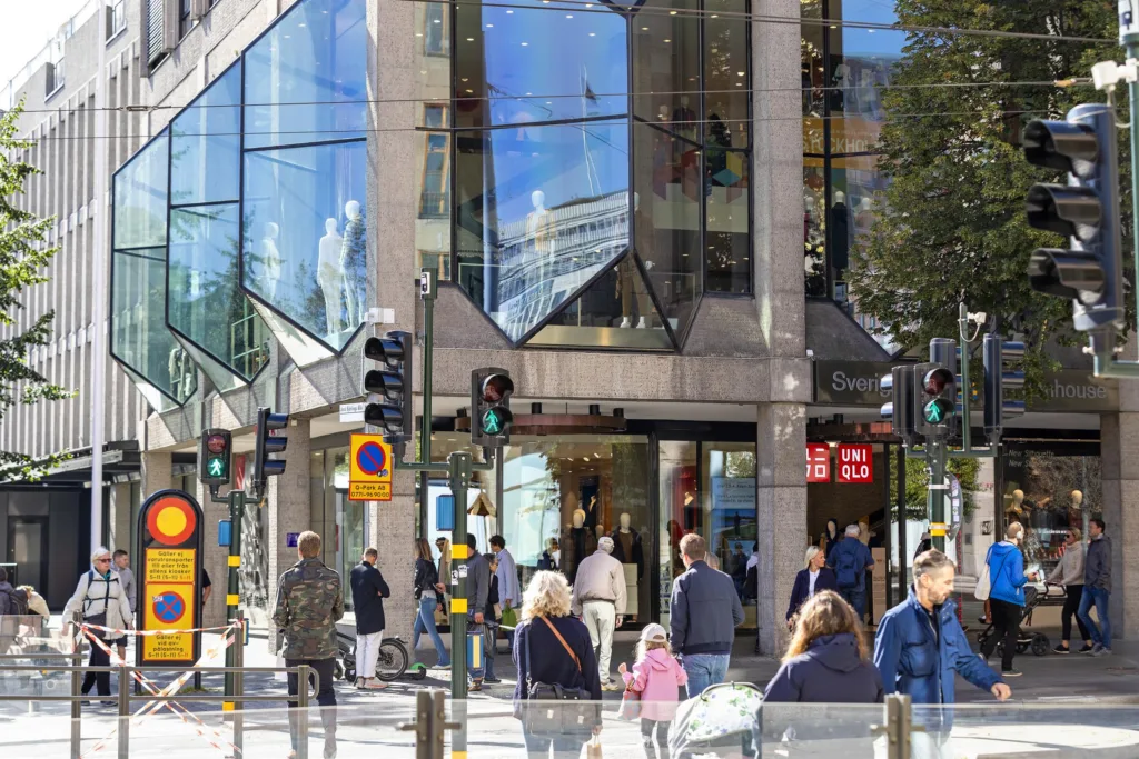 2. Gågata framför en modern byggnad med glasfasad, människor korsar vägen vid trafikljusen.