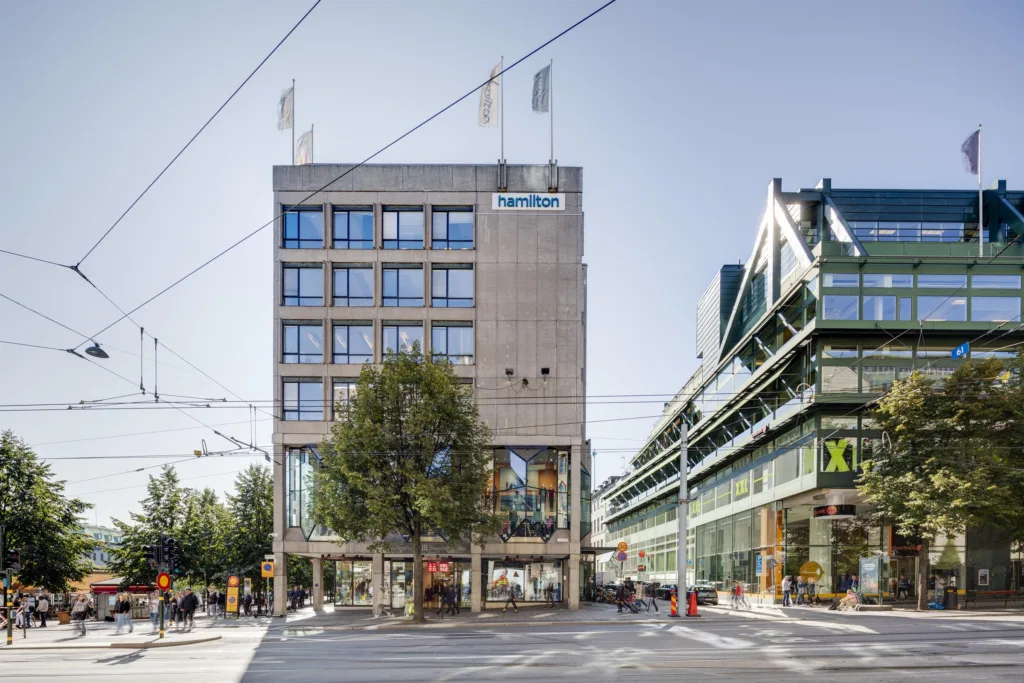 4. Byggnad med stora fönster och flaggor på taket samt en grön struktur till höger, människor rör sig på gatan.