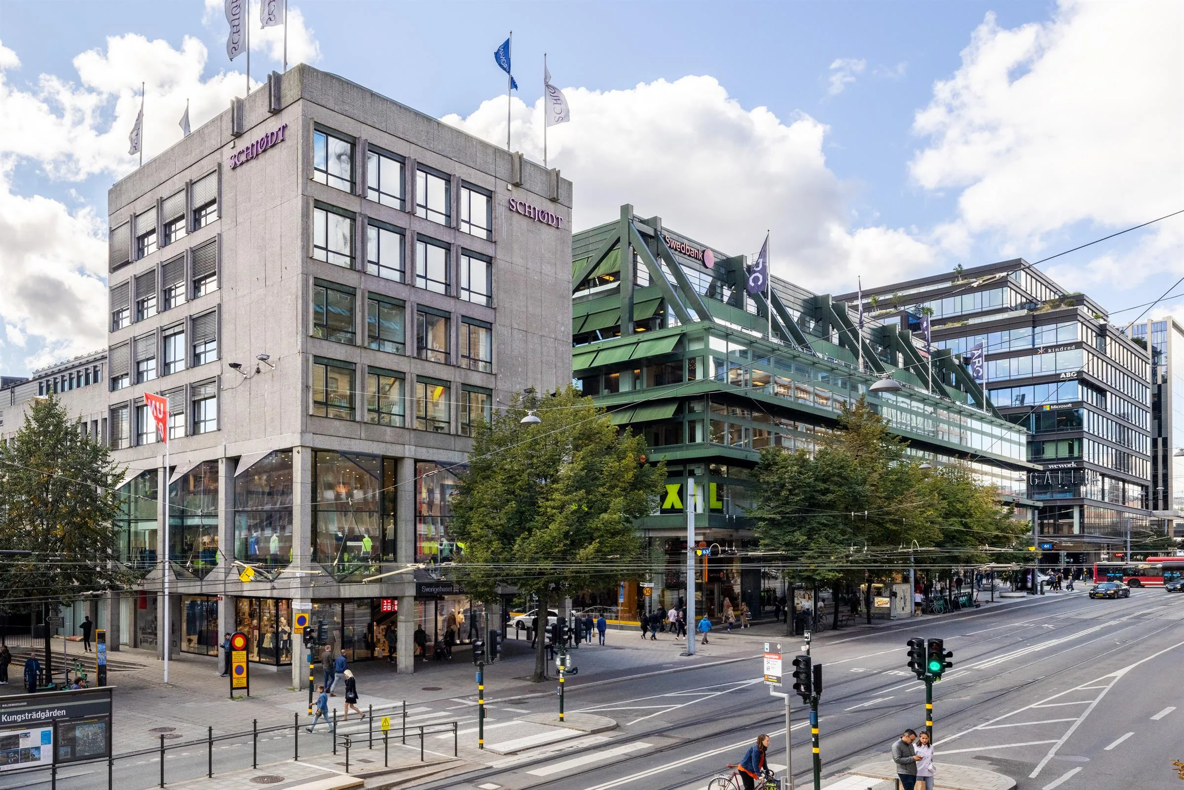 3. Hörnvy av en byggnad med flaggor på taket och grön struktur bredvid, trafik och människor på gatan.