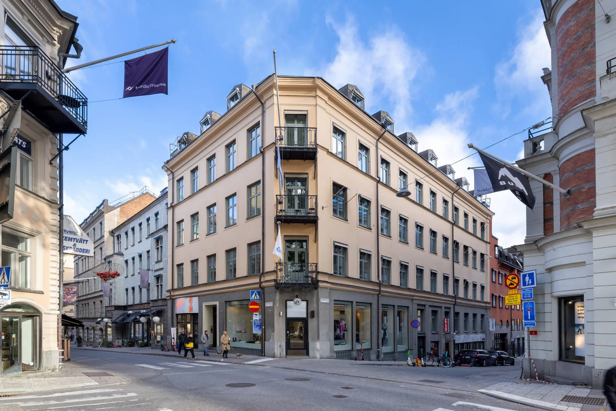 En beige hörnbyggnad med flera våningar och balkonger, omgiven av stadsgator med flaggor.