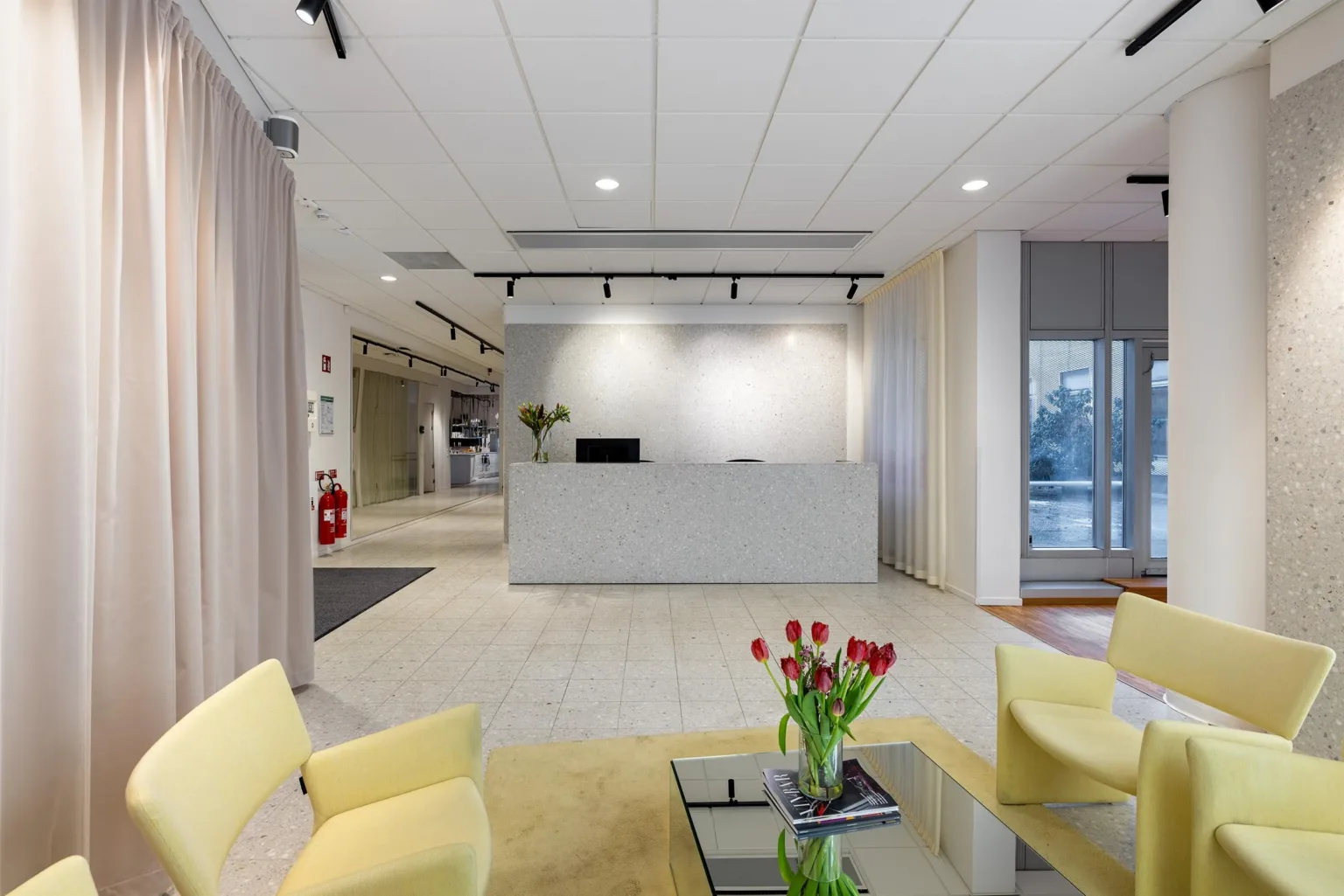 En reception med ett vitt receptionsbord, gula stolar, och en vas med röda tulpaner på glasbordet.