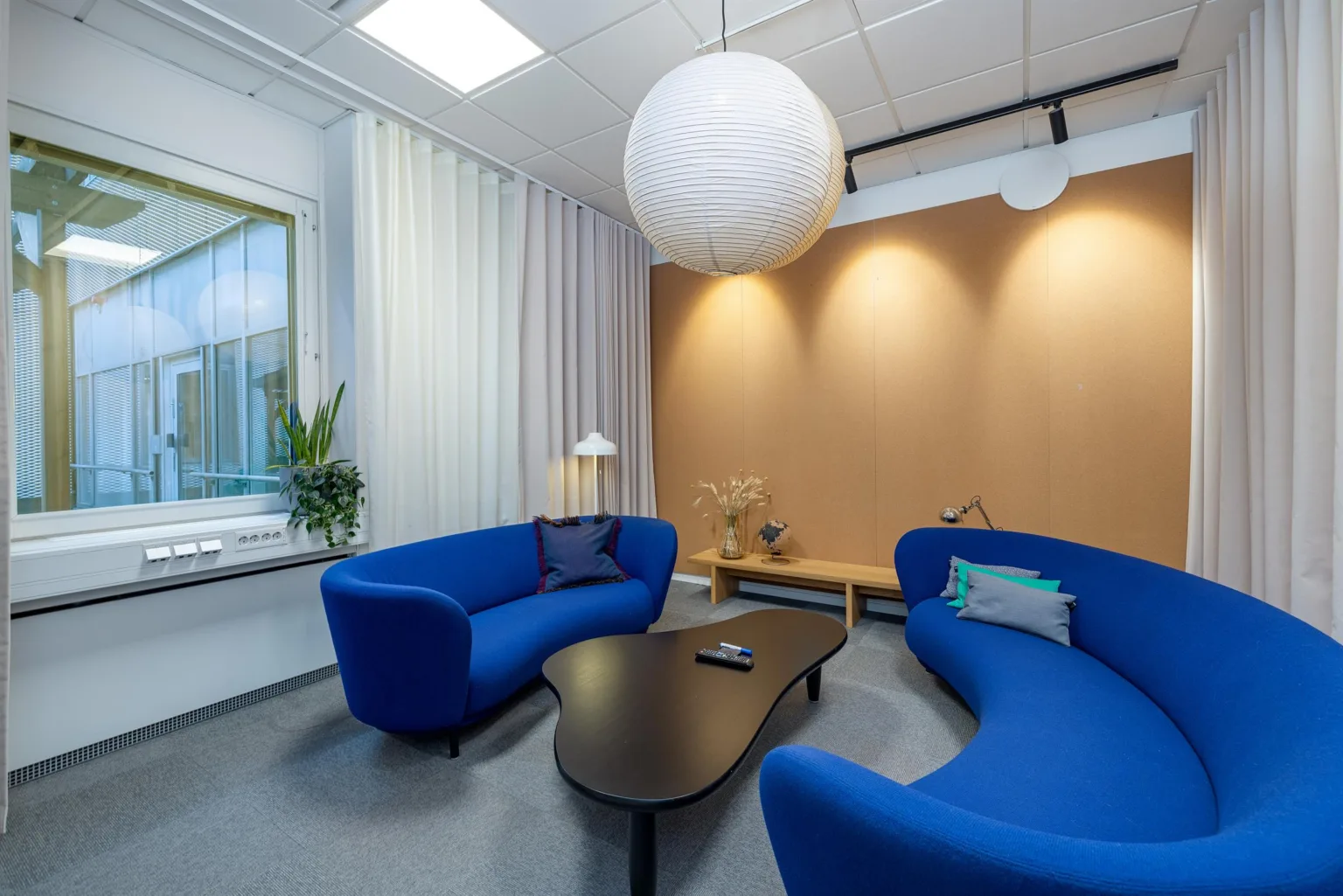 Ett rum med två blå soffor, ett mörkt soffbord, och en stor vit hängande lampa; beige vägg i bakgrunden.