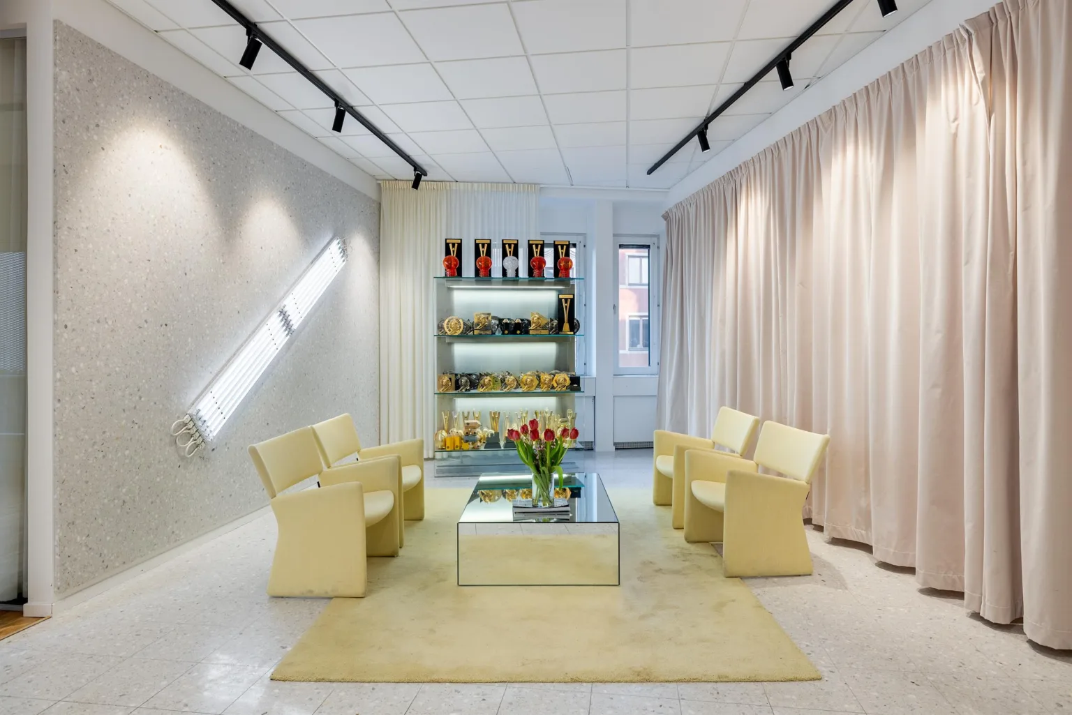 Modern lounge med fyra gula fåtöljer runt ett glasbord, hyllor i bakgrunden.