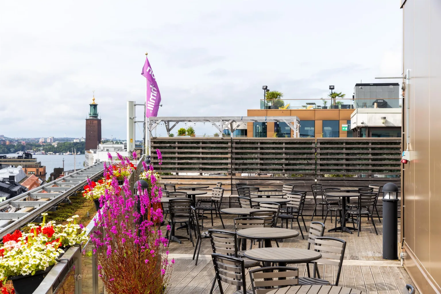 En takterrass med flera små bord och stolar, rosa blommor och utsikt över stadens byggnader och vatten.