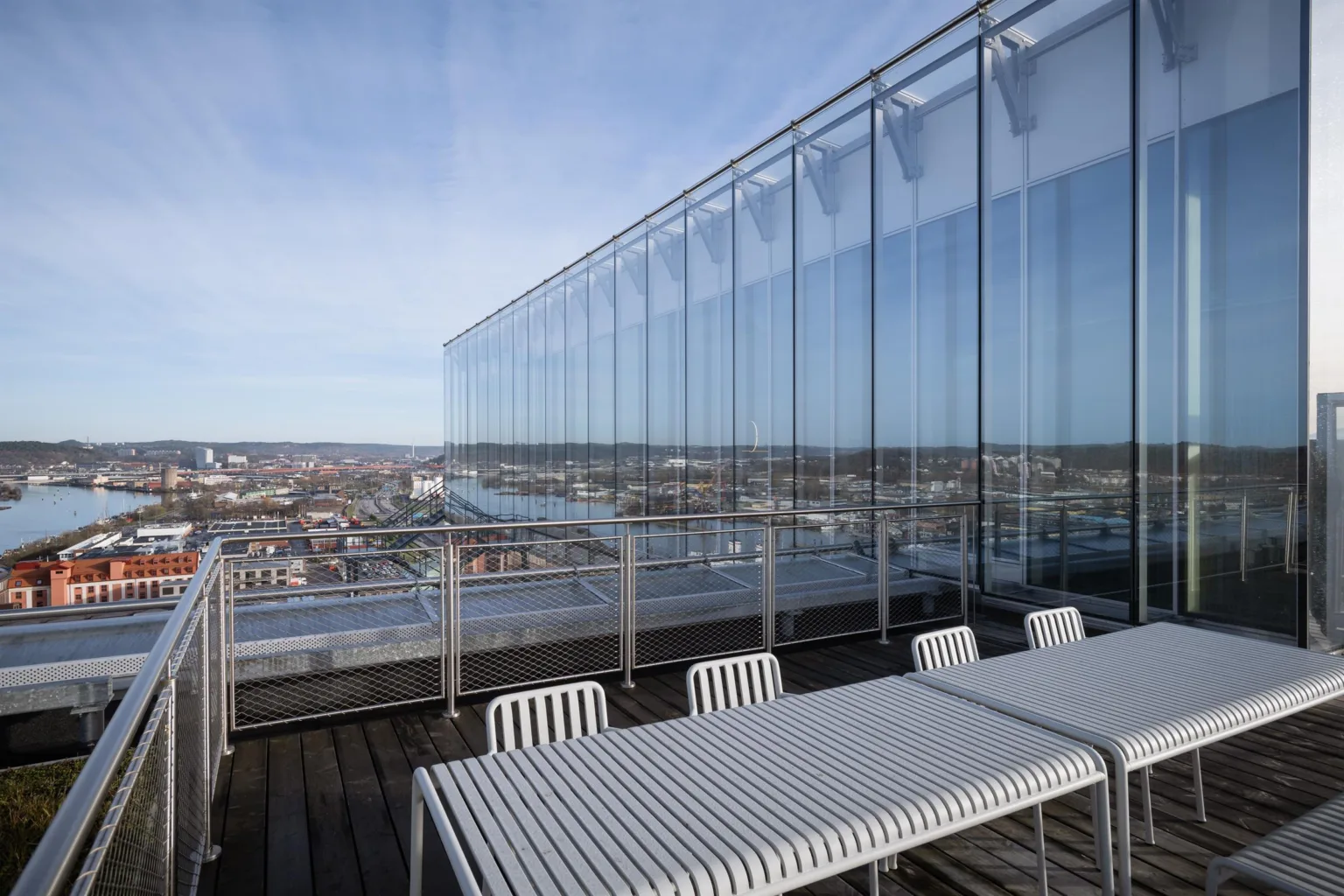 1. En takterrass med vita bord och stolar, omgiven av glasräcken och utsikt över en stad.