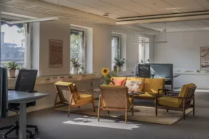 Ett kontorsrum med flera sittplatser, gula soffor, fåtöljer, ett glasbord med en solrosvas och flera fönster.
