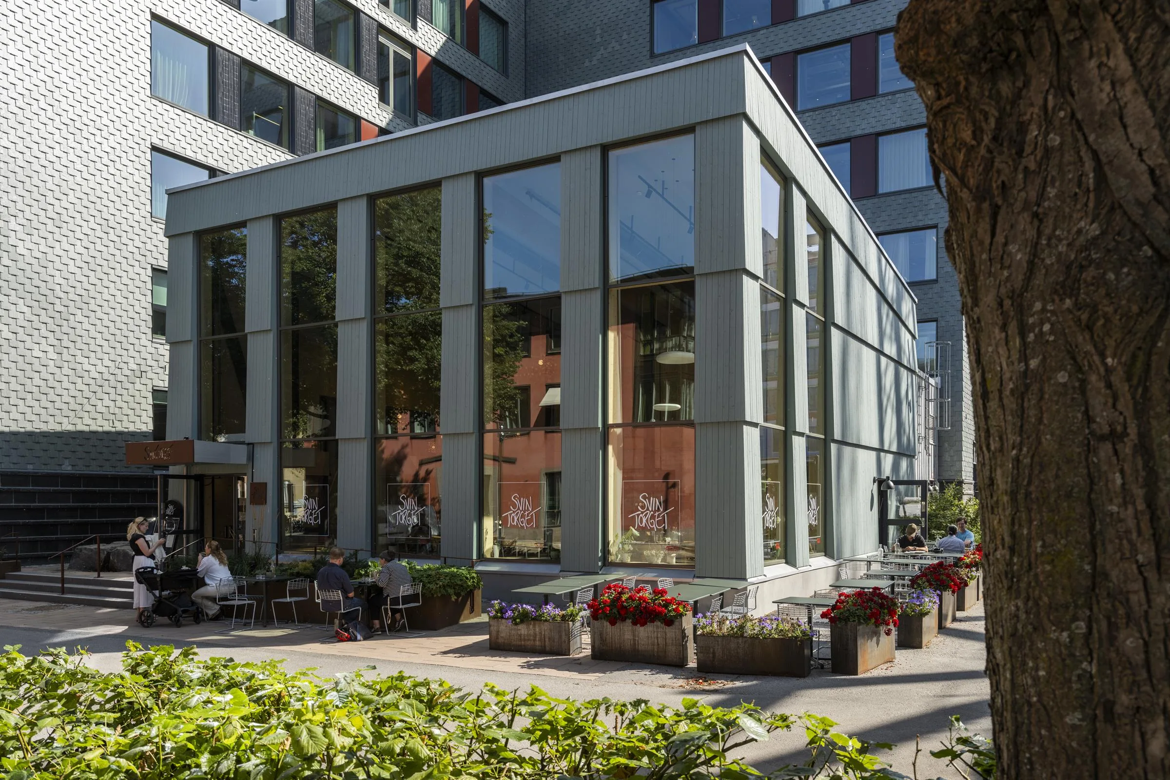 En modern byggnad med stora glasfönster, omgiven av grönska och blomsterdekorationer.