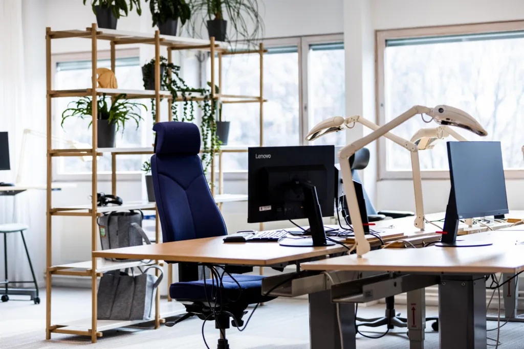 1. Kontorsmiljö med skrivbord, blå kontorsstol, datorskärm och hyllor med växter vid stora fönster.