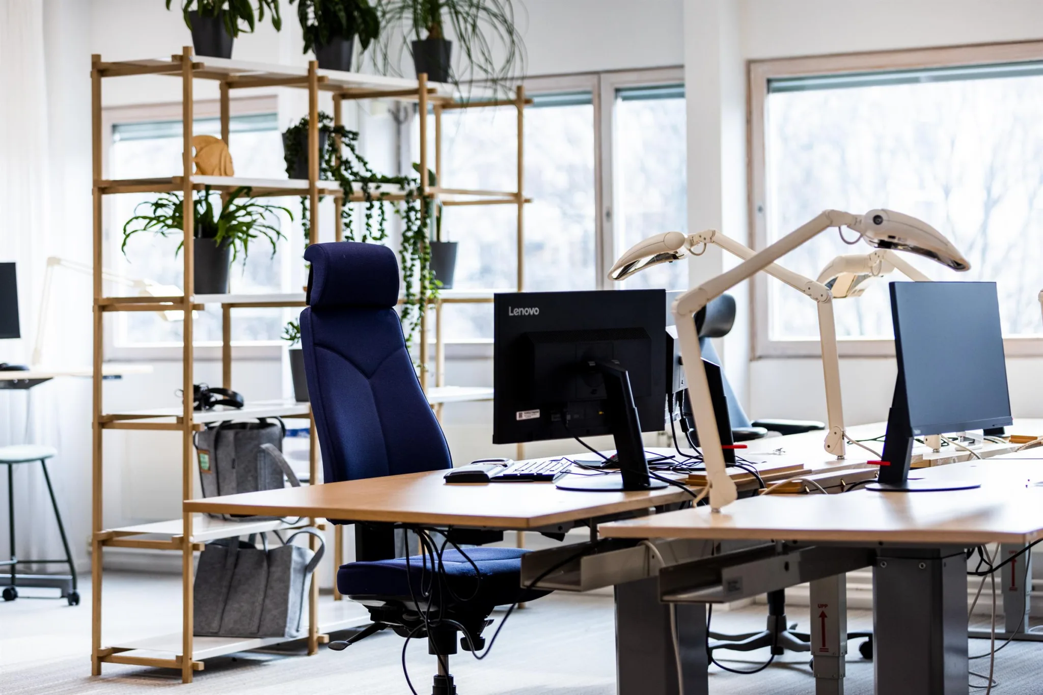 1. Kontorsmiljö med skrivbord, blå kontorsstol, datorskärm och hyllor med växter vid stora fönster.