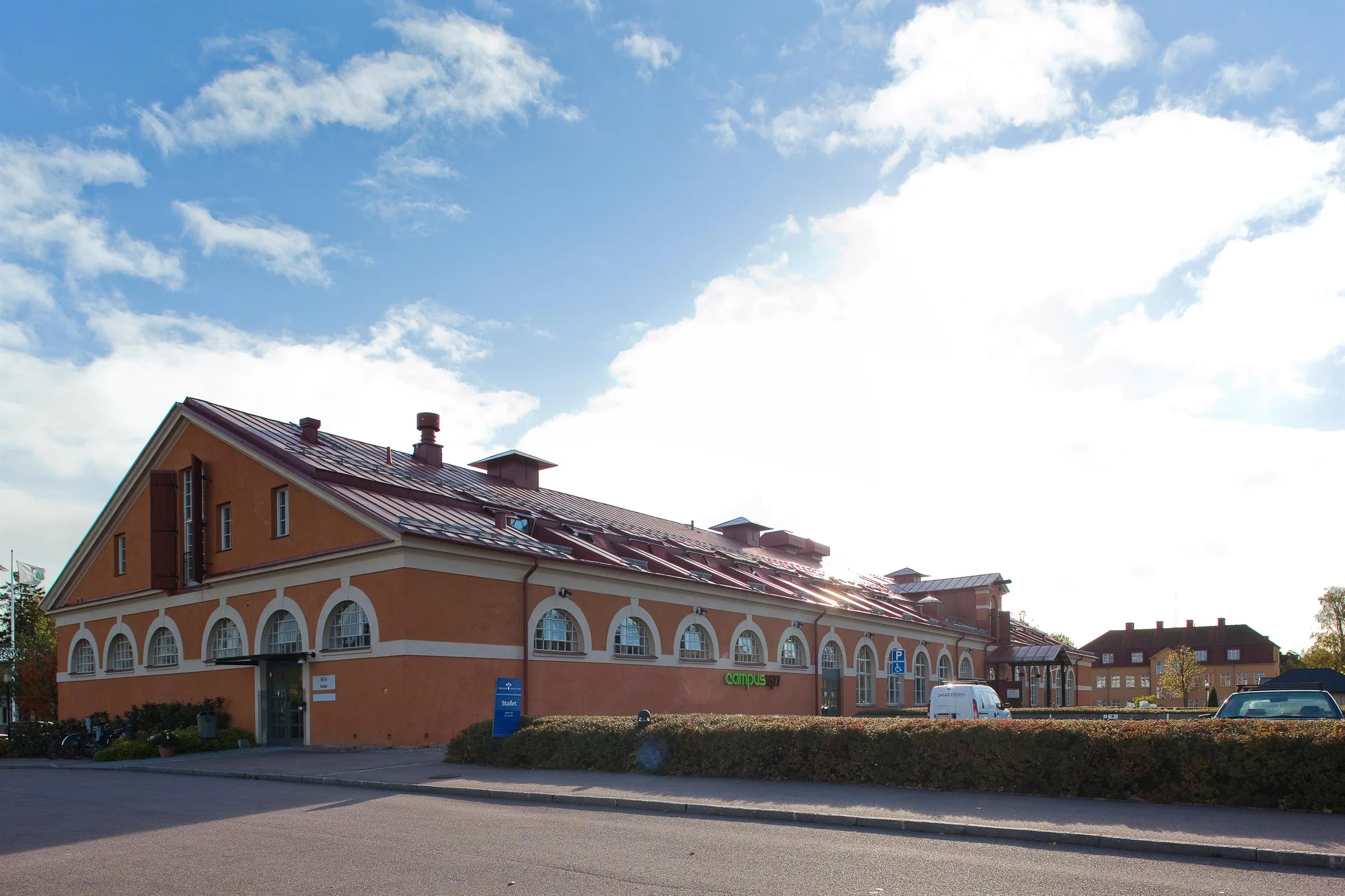 En lång, gul-orange byggnad med bågformade fönster och röda tak, med en parkering framför.
