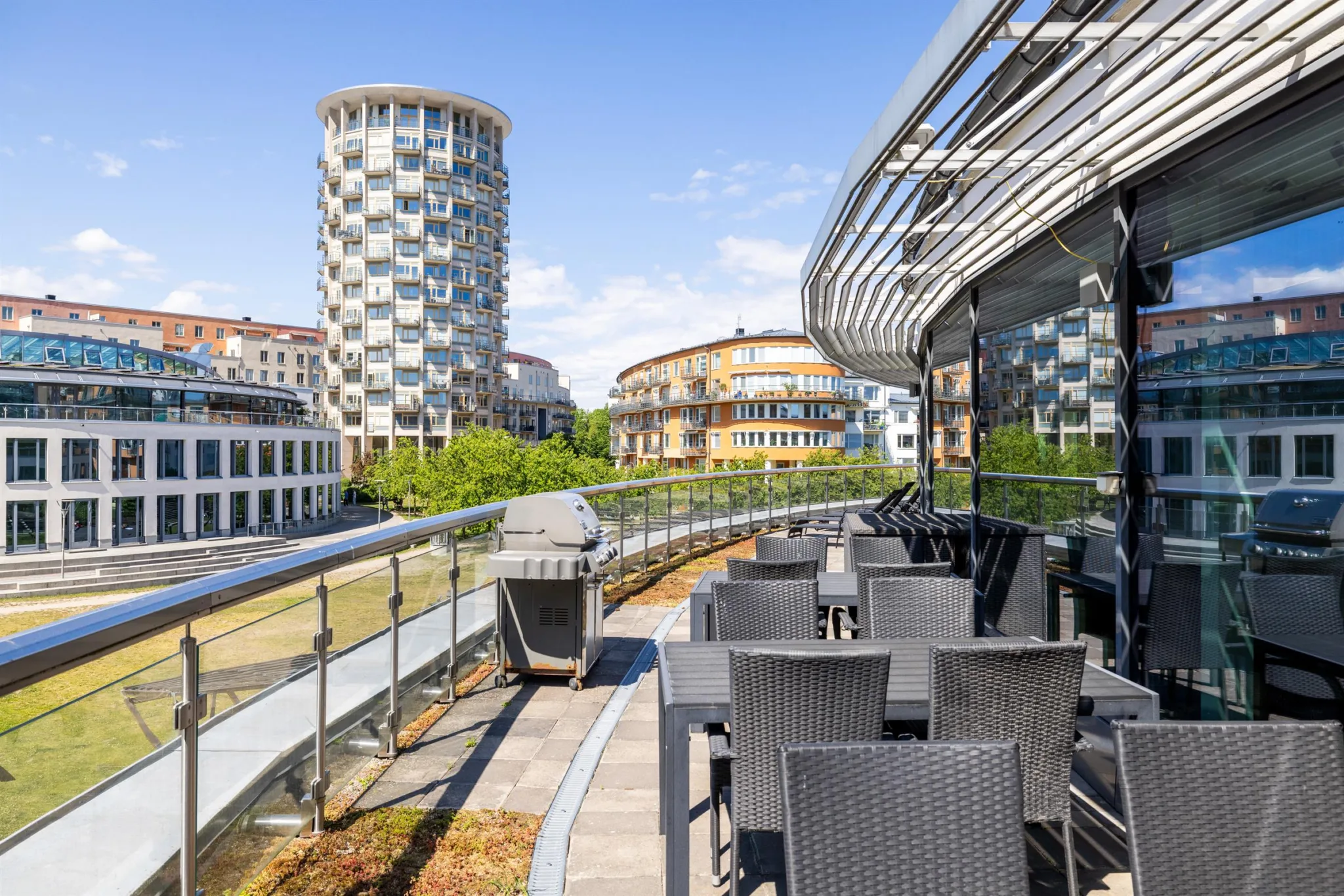 En utomhusterrass med svarta utemöbler, glasräcke och utsikt över moderna byggnader och grönområden.