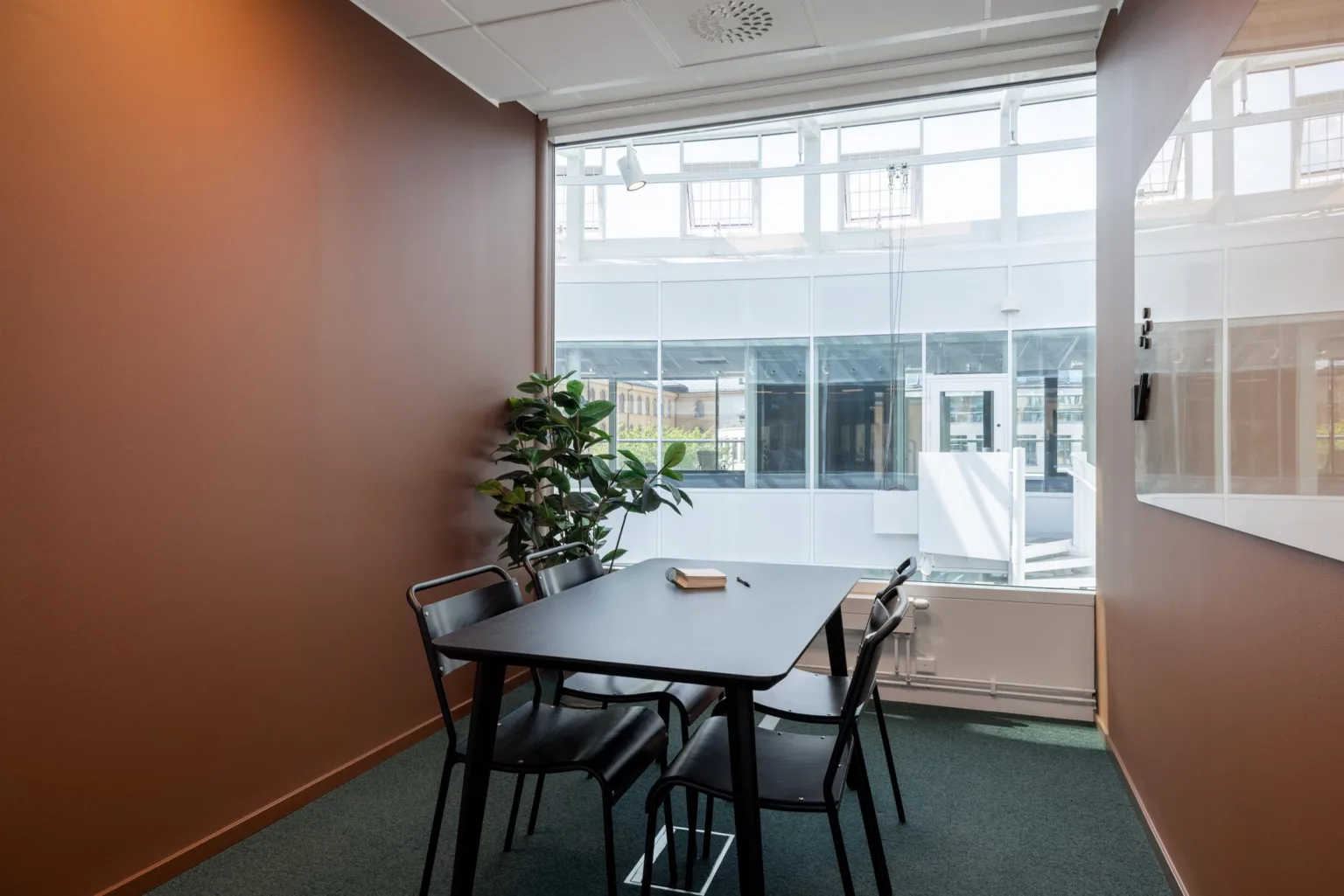 Litet mötesrum med bord, fyra stolar, grön växt och stort fönster med utsikt mot innergården.