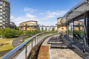 En terrass med glasräcke, solstolar och utsikt över en park med gröna omgivningar och flerbostadshus i bakgrunden.
