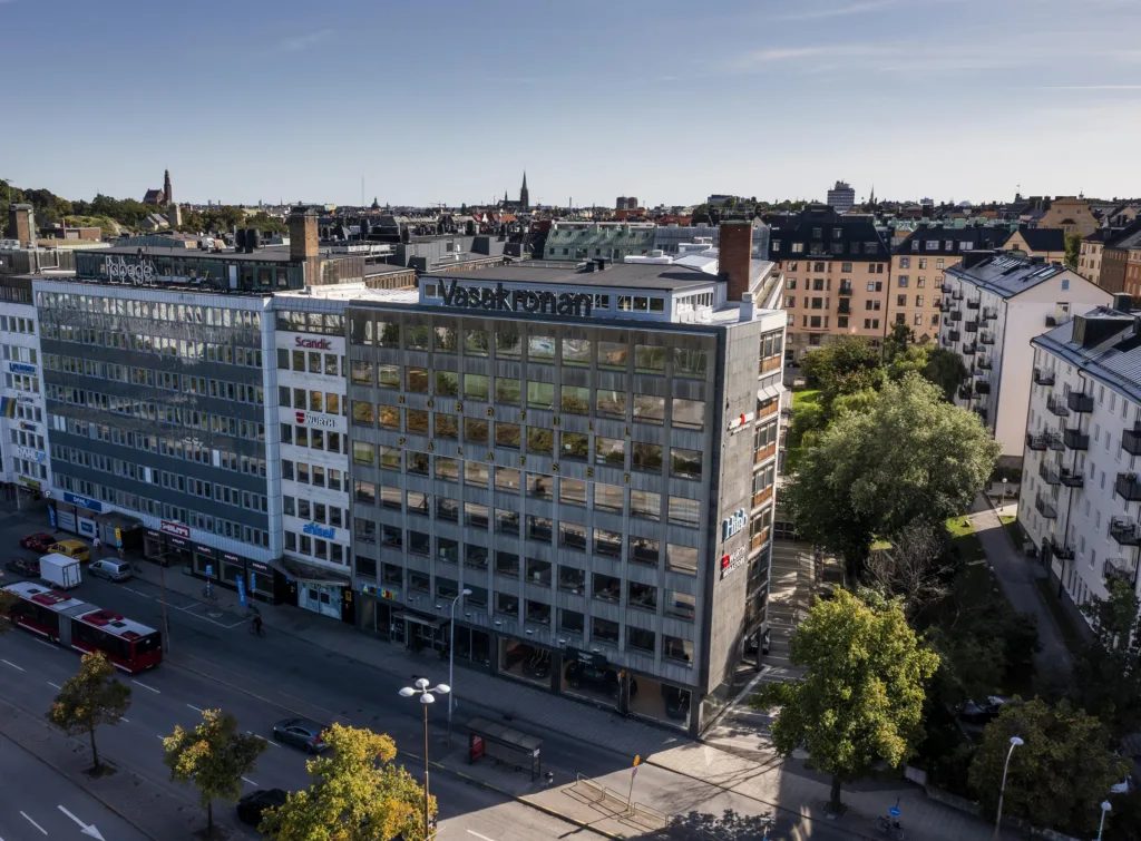 En byggnad med modern arkitektur, omgiven av grönska och stadsmiljö.