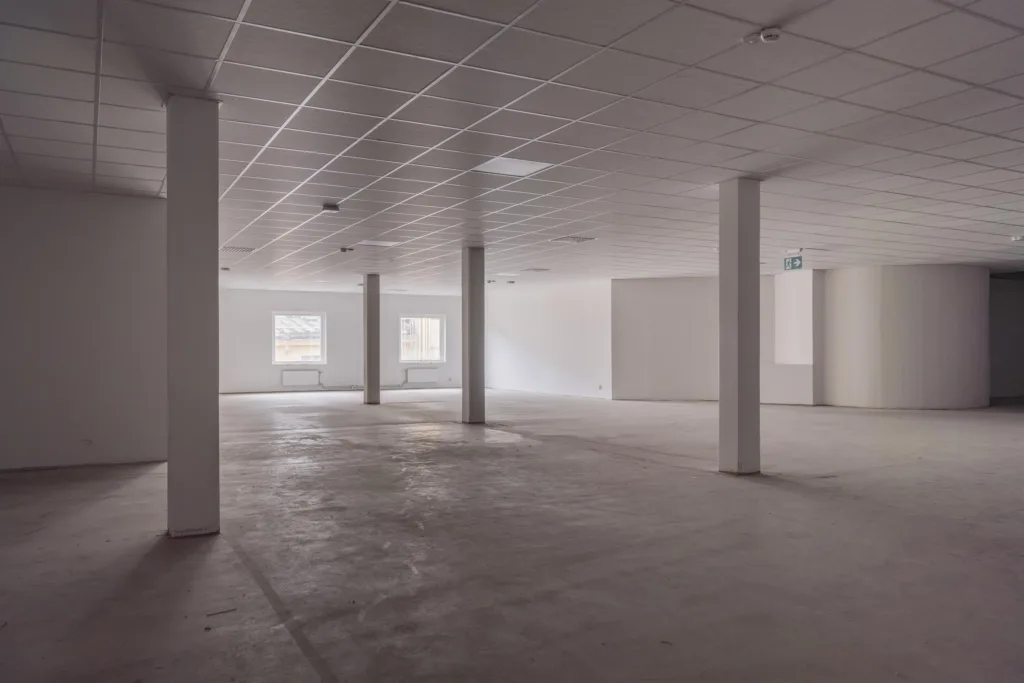 Ett öppet utrymme med pelare, vitmålade väggar och tak, och betonggolv. Fönster syns i bakgrunden.