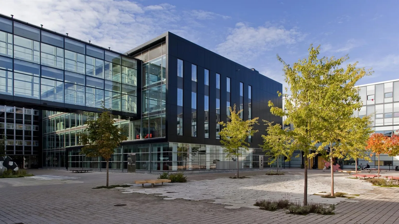 5. En modern byggnad med glasfasad och mörka väggar, omgiven av träd och en stenbelagd gårdsplan.