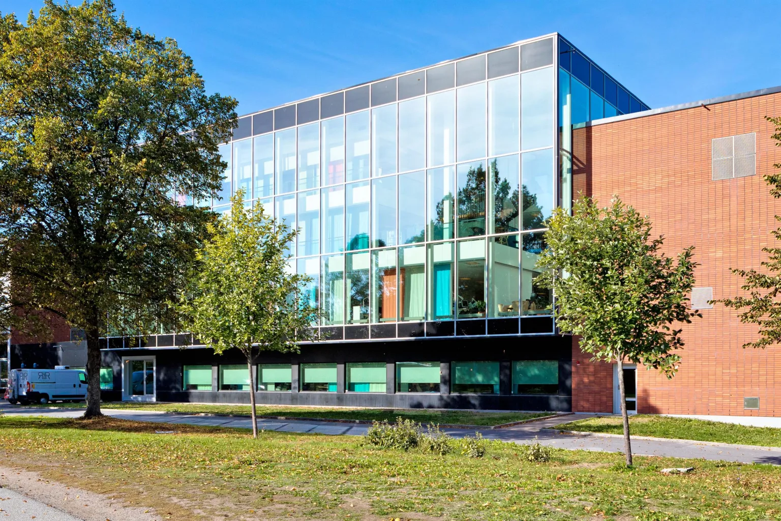 En modern byggnad med stora glasfasader omgiven av träd och grönska.