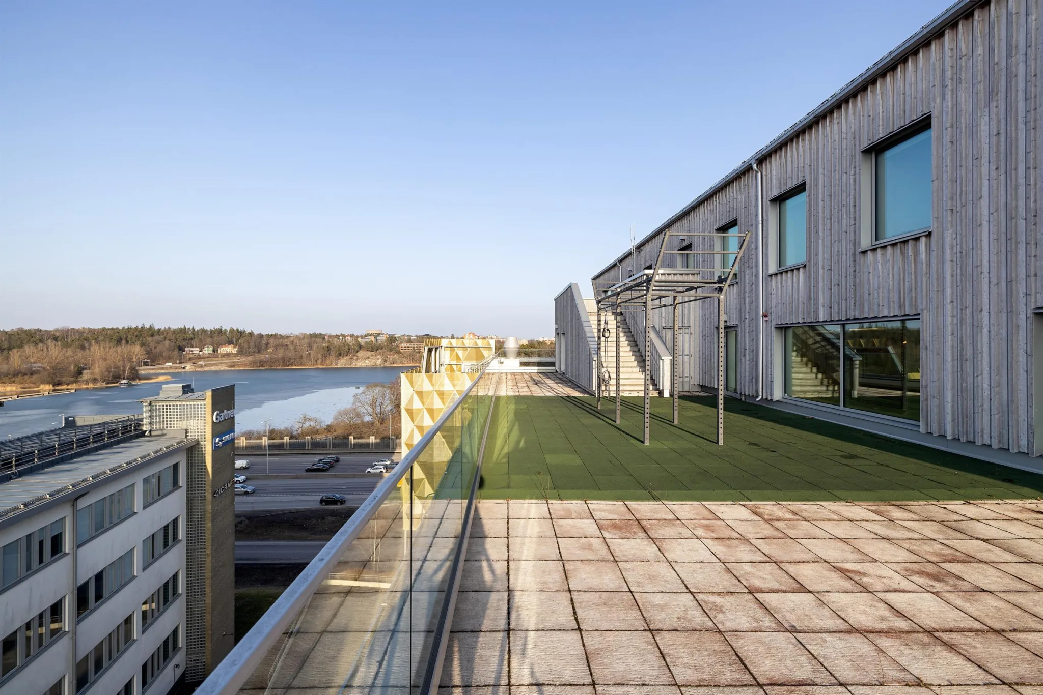 En takterrass med glasräcke, grön konstgräsmatta, metallställningar och utsikt över byggnader och landskap.