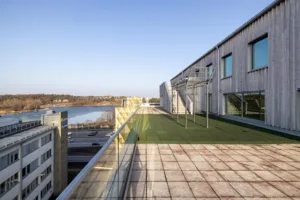 En takterrass med glasräcke, grön konstgräsmatta, metallställningar och utsikt över byggnader och landskap.
