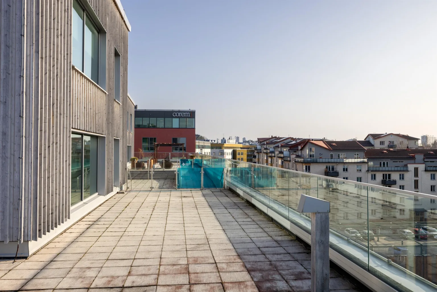 En terrass med glasräcke, utsikt över stadsmiljö och en träklädd byggnad till vänster.