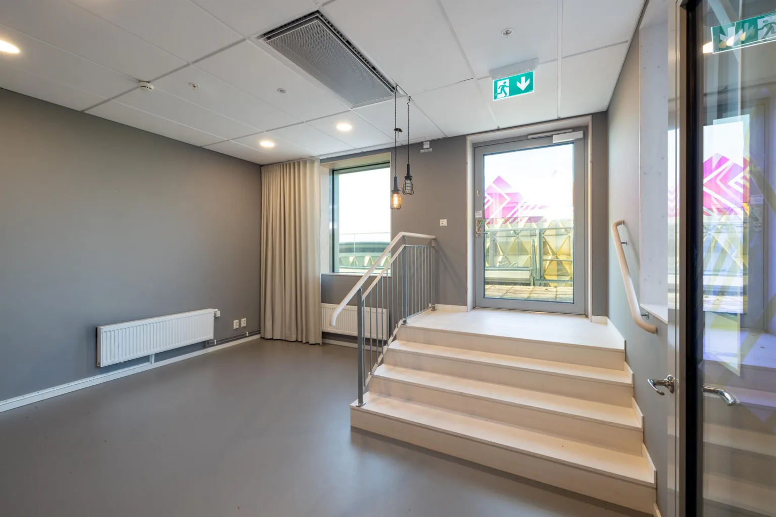 5. Ett rum med grå golv, trappa, dörr till en balkong och ett fönster. Exit-skylt ovanför dörren.
