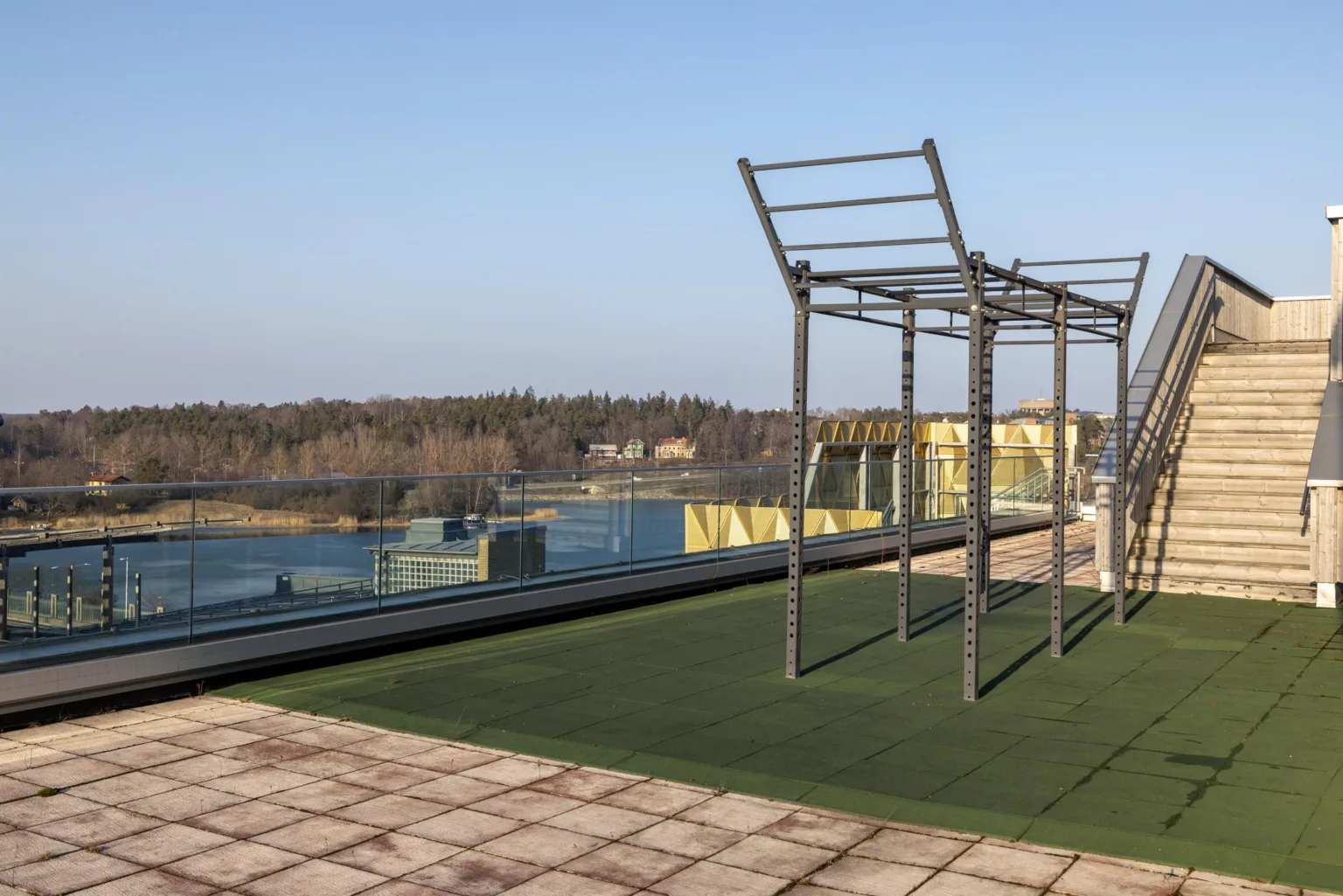 En takterrass med grön konstgräsmatta och metallställningar, utsikt över ett skogsområde och vatten.