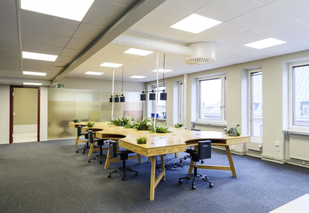 Ett modernt kontor med ett långt träbord, svarta kontorsstolar och växter, omgivna av stora fönster.