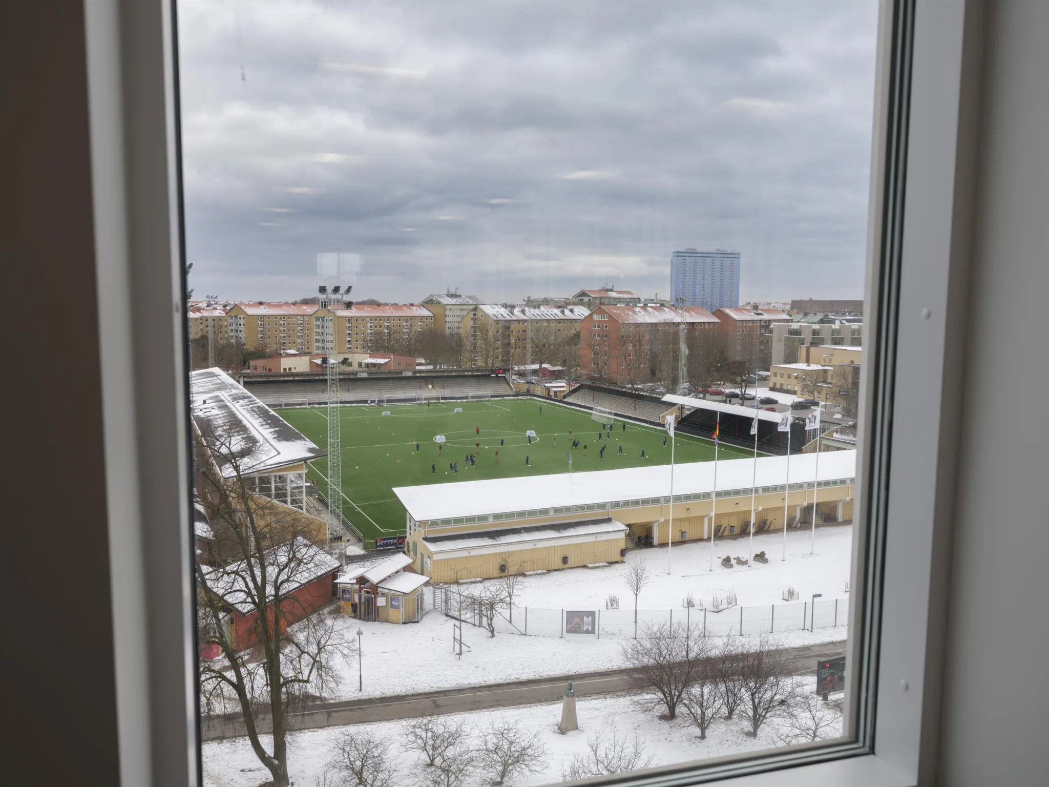 Utsikt från ett fönster över en snötäckt idrottsplan och omgivande byggnader.