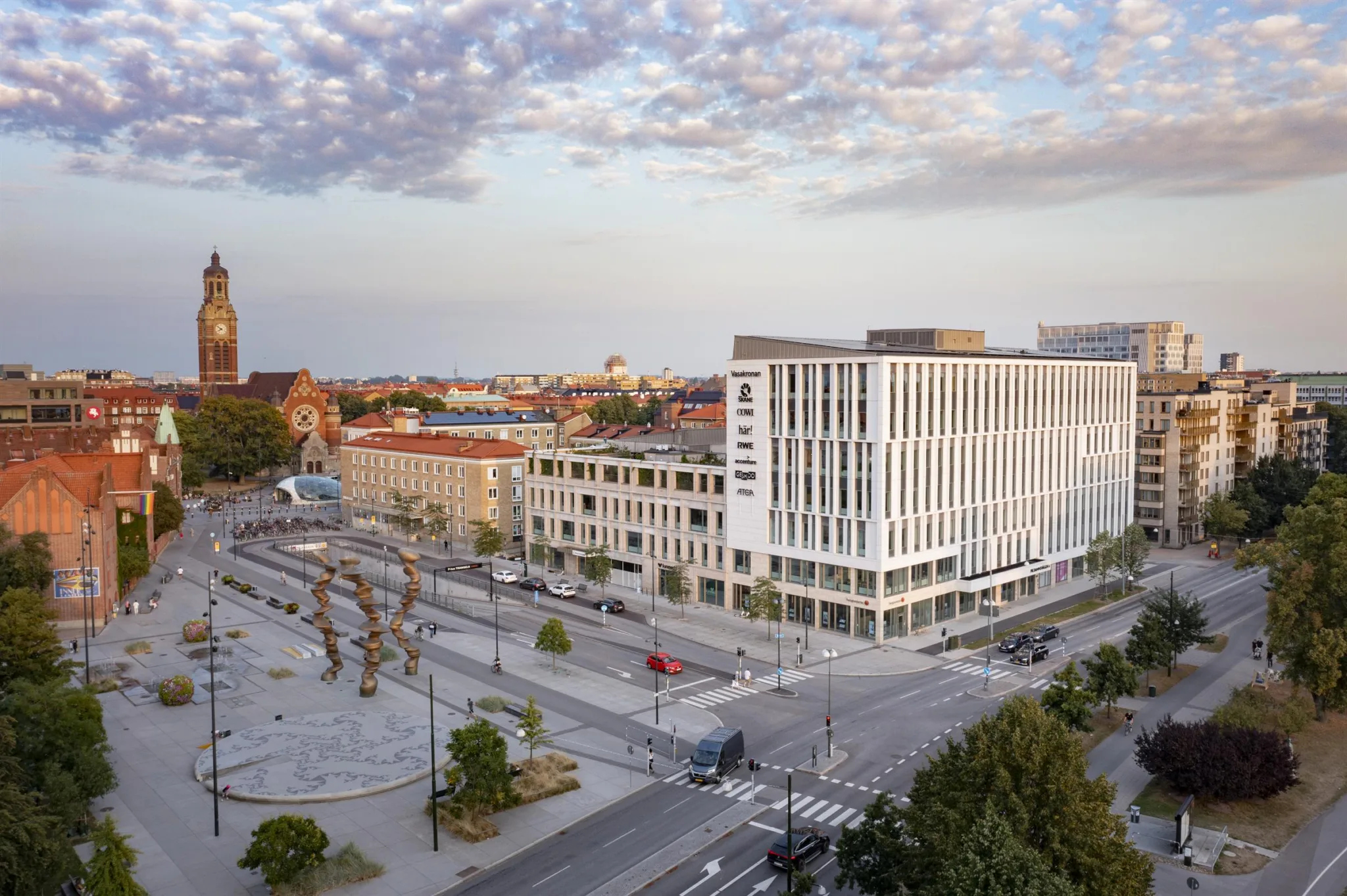 En vy över en stadsdel med moderna byggnader, ett öppet torg med träd och bilar på gatorna under en delvis molnig himmel.