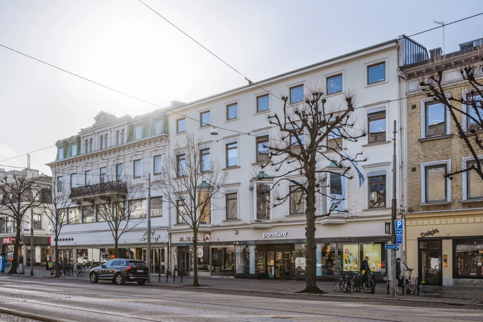 En vit byggnad med tre våningar och flera fönster längs en gata med parkerade bilar och träd.