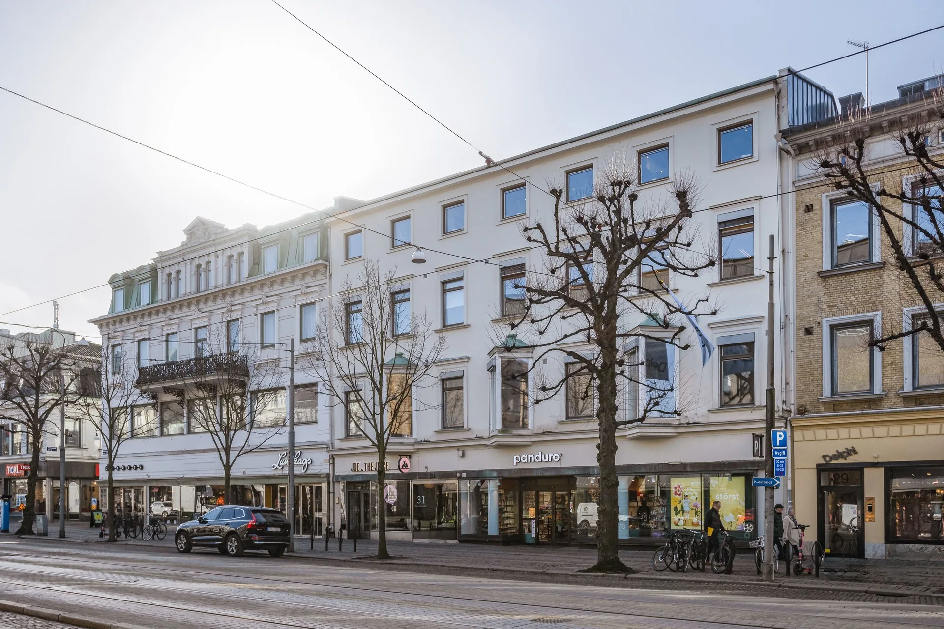 En vit byggnad med tre våningar och flera fönster längs en gata med parkerade bilar och träd.
