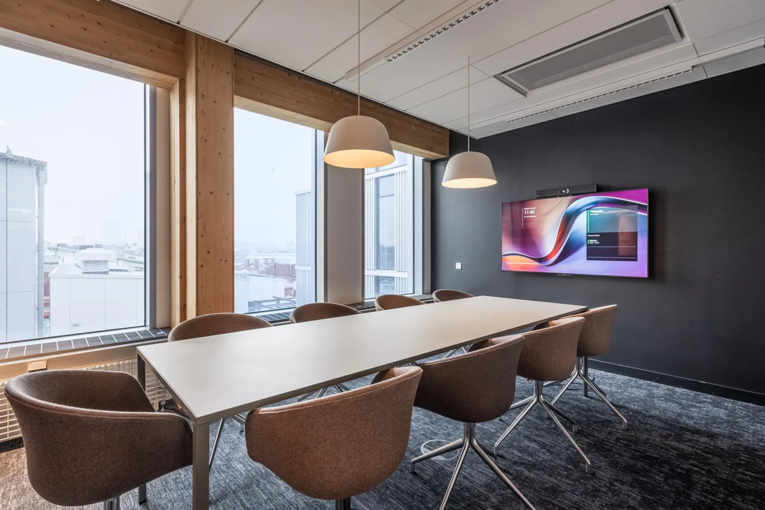 1. Ett modernt konferensrum med ett långt bord, åtta stolar, taklampor och en stor väggmonterad skärm.