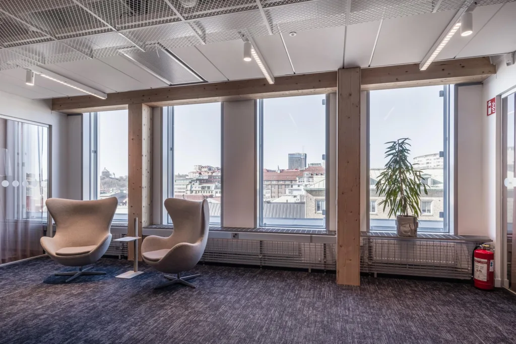 1. Två moderna fåtöljer bredvid stora fönster med utsikt över en stadsmiljö, samt en grön växt i ett hörn.
