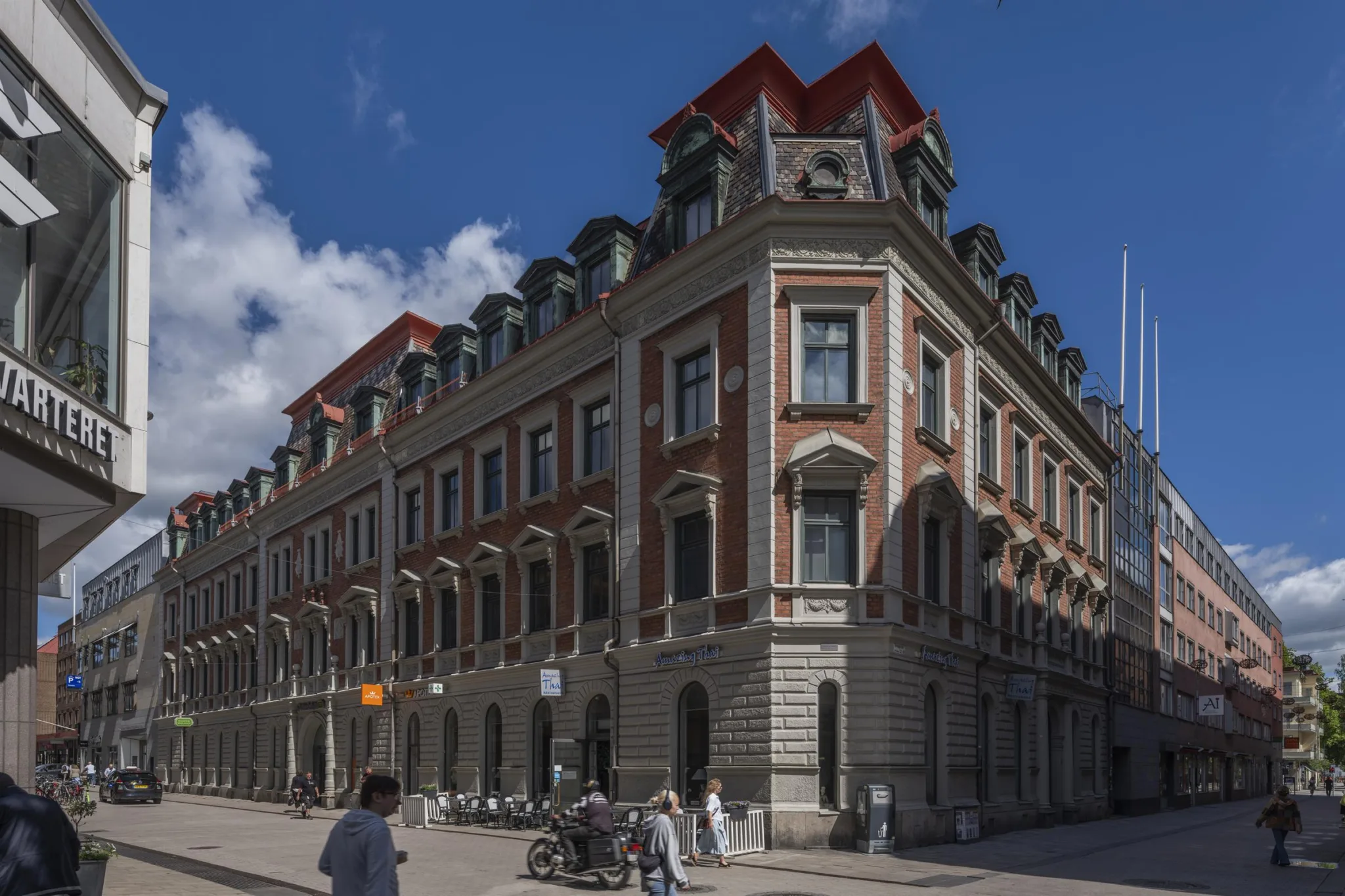 En stor byggnad med röd taknock och detaljrik arkitektur, människor rör sig och sitter på bänkar under blå himmel.