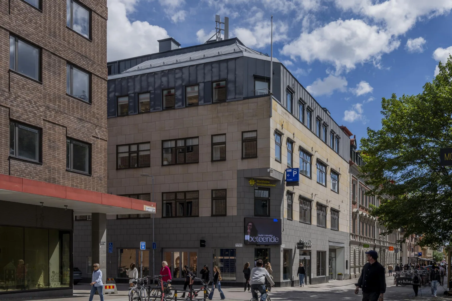 En beige byggnad med stora fönster, människor går på trottoaren, och himlen är blå med vita moln.