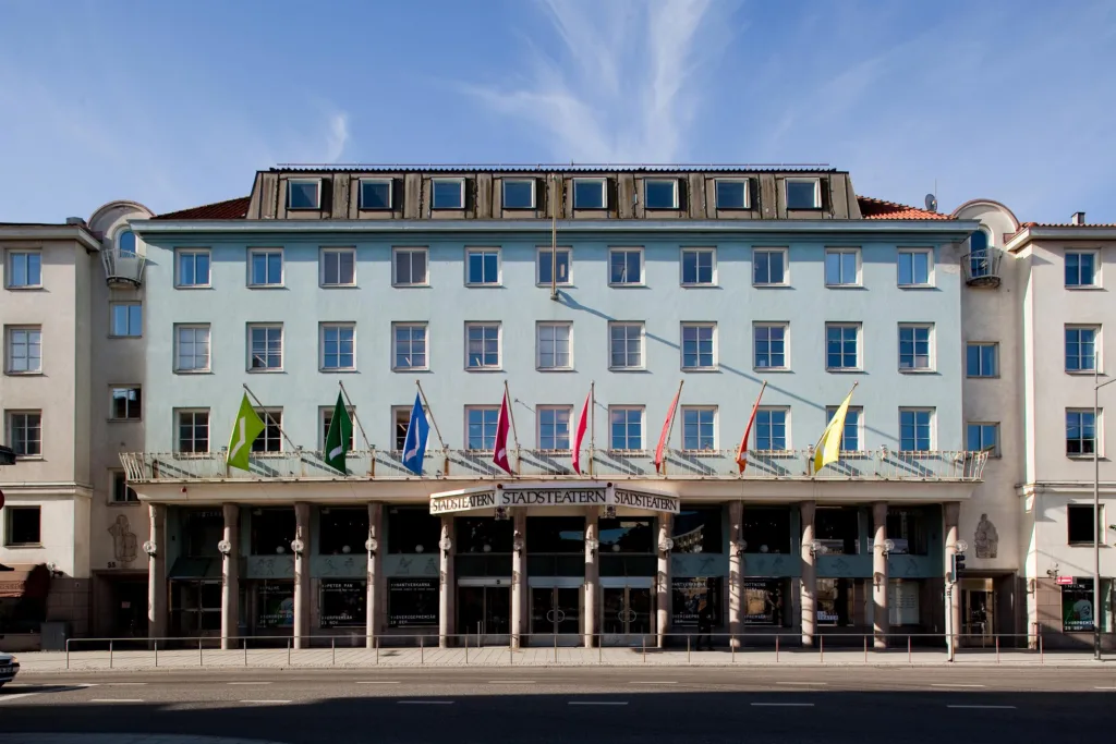 En ljusblå byggnad med flera flaggor vid entrén; himlen är klar med några moln.
