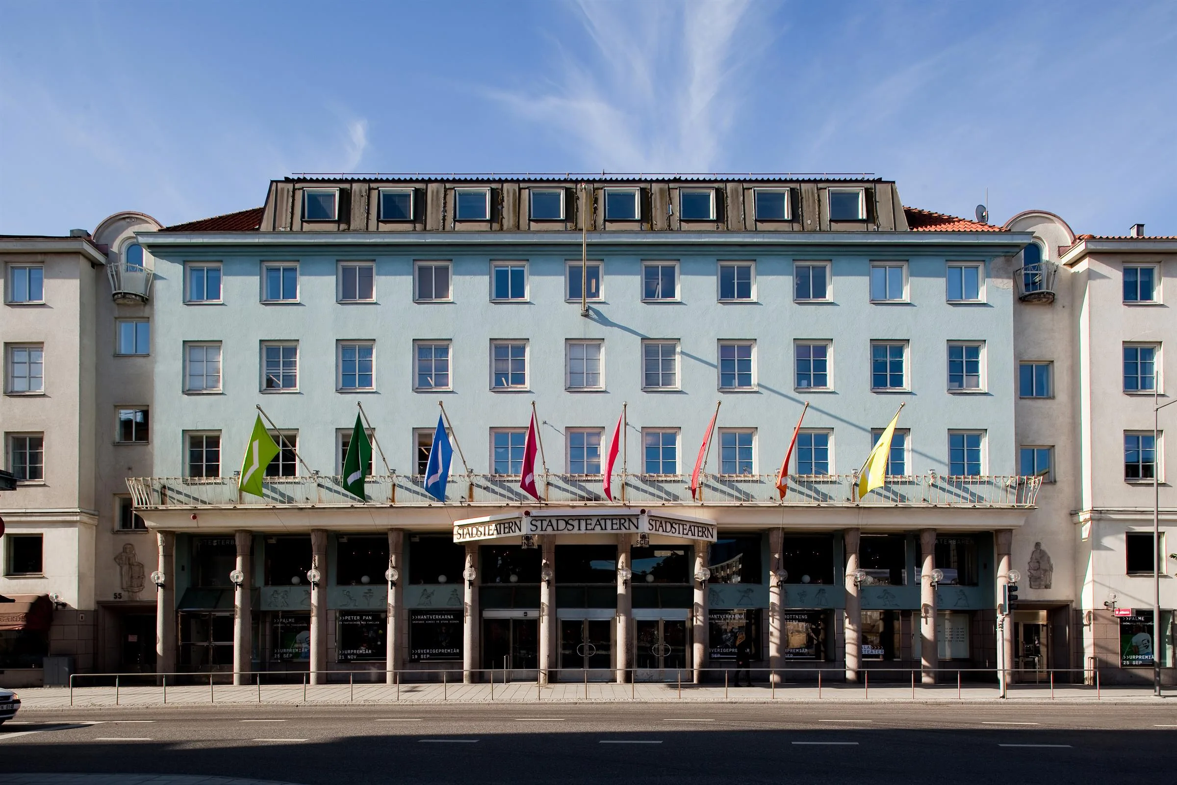 En ljusblå byggnad med flera flaggor vid entrén; himlen är klar med några moln.