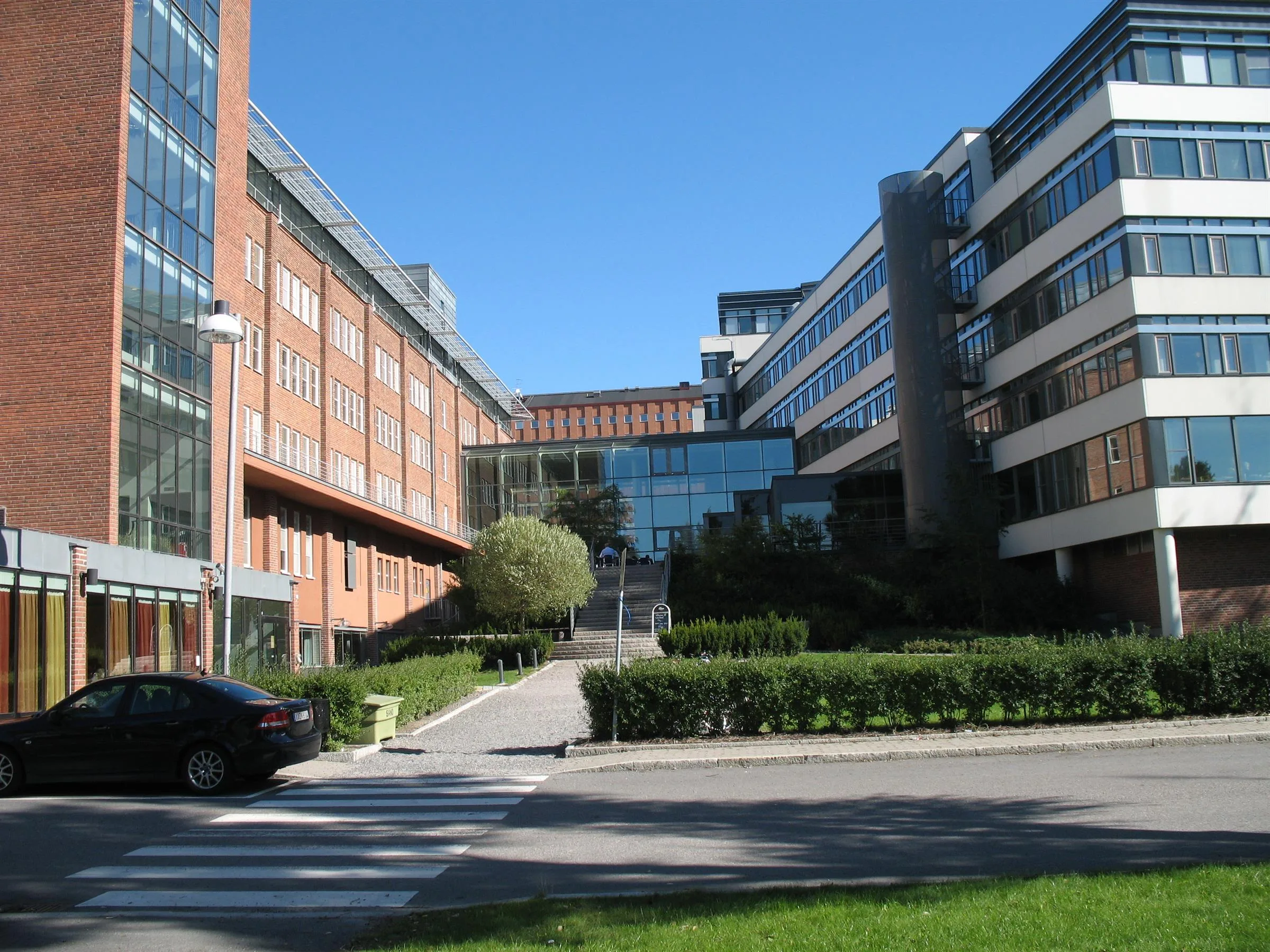 En byggnad med moderna fasader, omgiven av gröna häckar och en gångväg under en klarblå himmel.