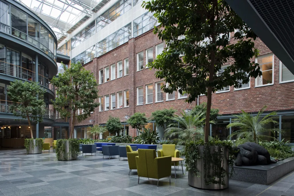 En innergård med gröna träd, färgglada sittmöbler och ett glastak ovanför.