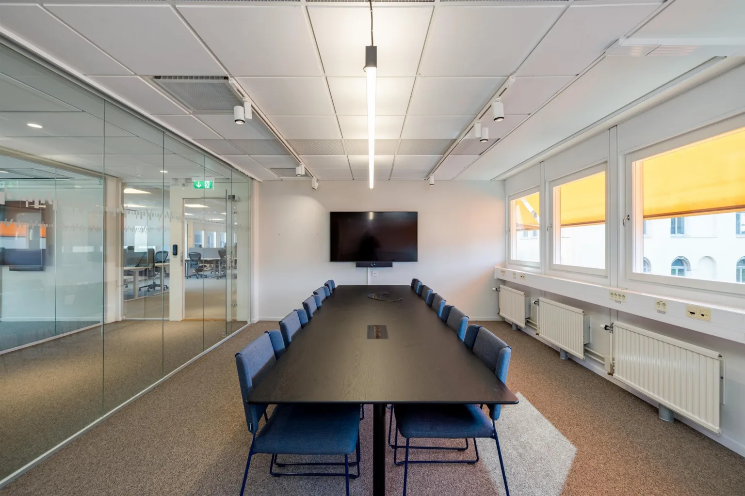 Ett rymligt konferensrum med långbord, blå stolar, en väggmonterad skärm och glasväggar mot en öppen kontorsyta.