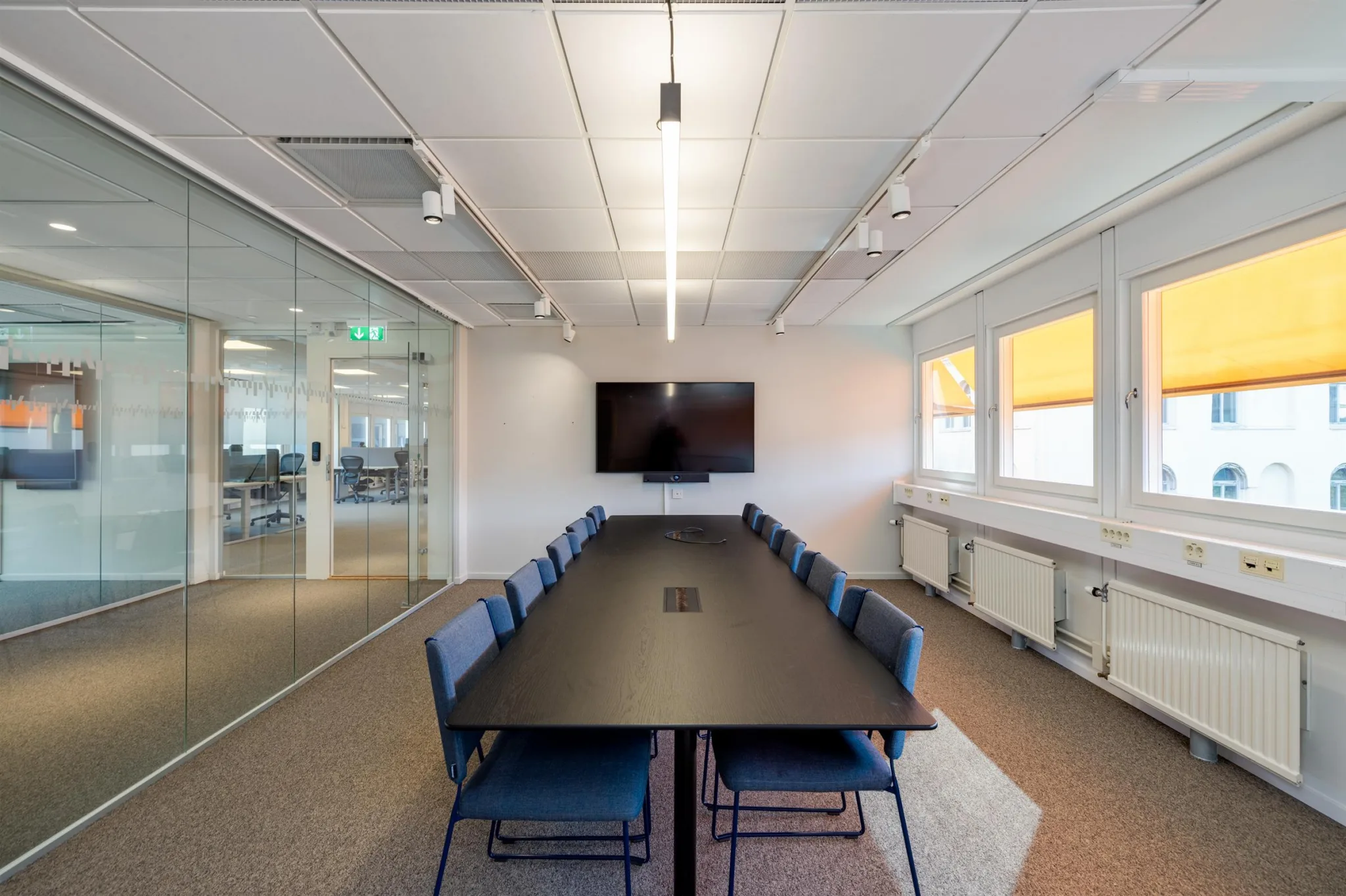 Ett rymligt konferensrum med långbord, blå stolar, en väggmonterad skärm och glasväggar mot en öppen kontorsyta.