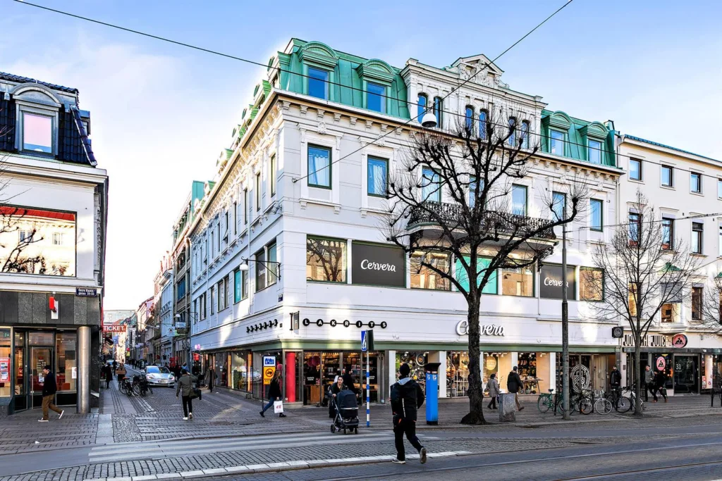 En hörnbyggnad med vit fasad och grönt tak, butikslokaler på bottenvåningen och människor som passerar på gatan.