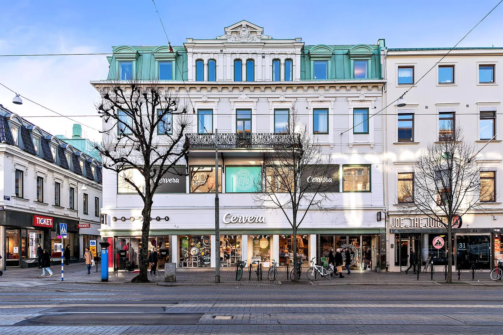 En vit byggnad med gröna tak och flera fönster, butikslokaler längst ner och människor på gatan.