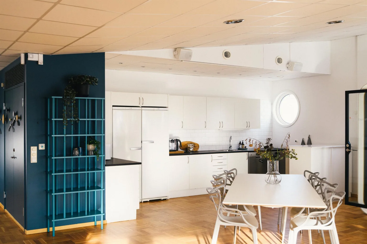 Ett öppet köksutrymme med vita skåp, en blå hylla och ett matbord med stolar.