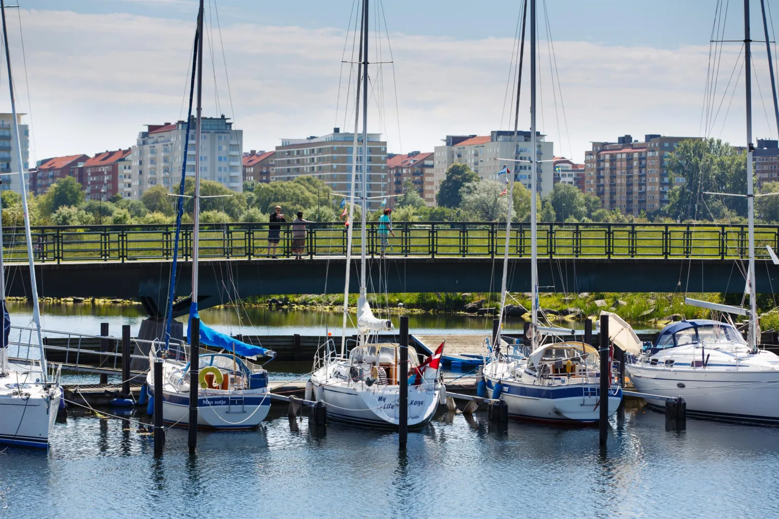 Små segelbåtar förtöjda vid en brygga, med en bro och byggnader i bakgrunden.