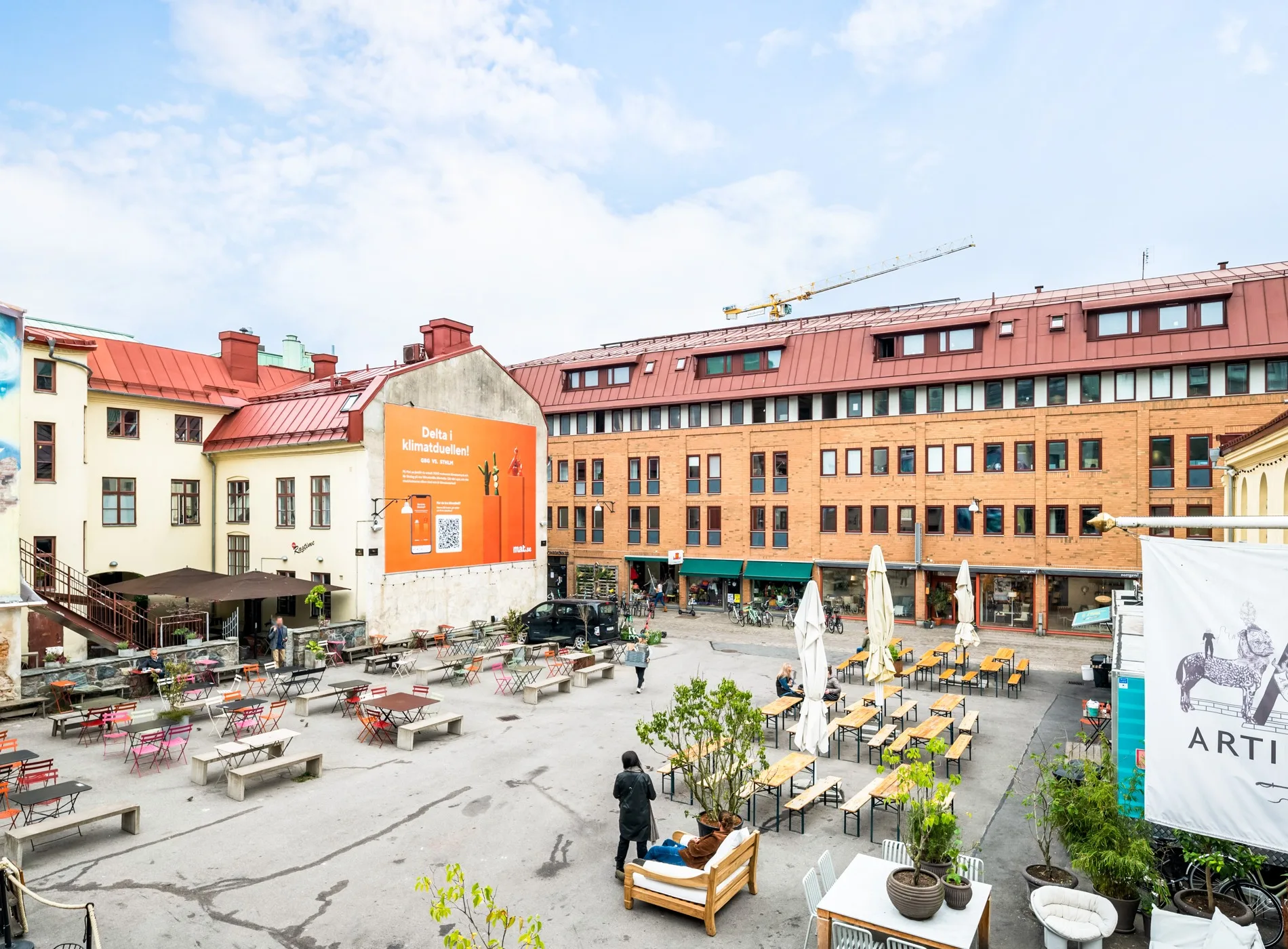 En öppen innergård med sittplatser, växter och parasoller, omgiven av röda tegelbyggnader.