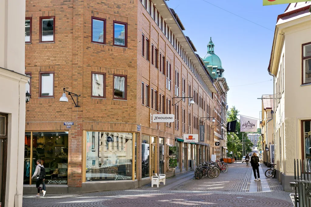 En gata med tegelbyggnader, butiker och cyklar; ett torn med grön kupol syns i bakgrunden.